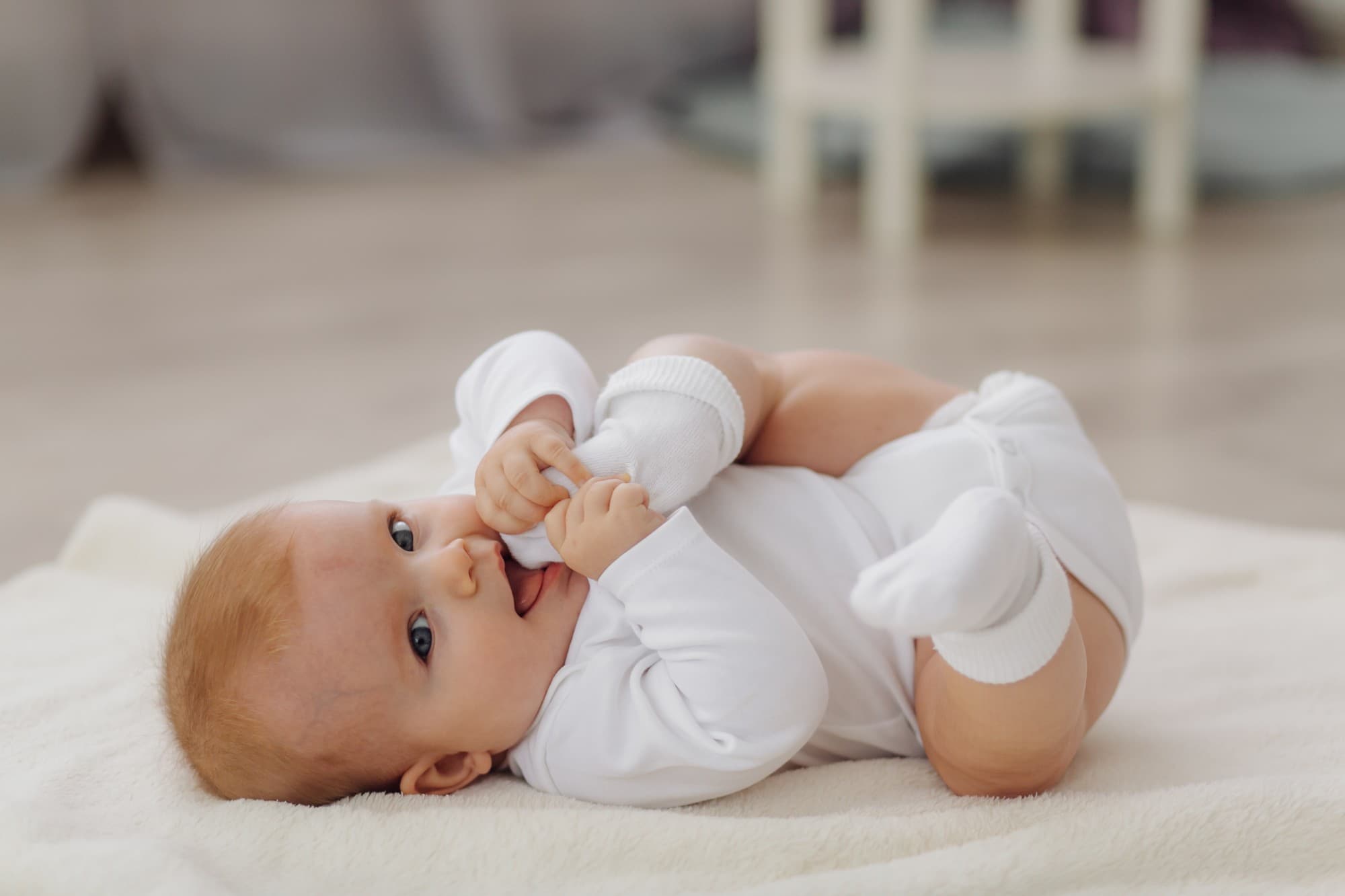 Bebê de body branco deitado sobre uma manta, segurando o pé e olhando para a câmera