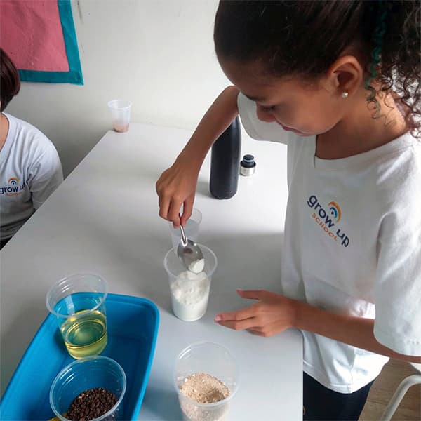 Aluna do fundamental I fazendo um experimento: coloca um pó branco no copo com uma colher, com óleo e grãos separados ao lado