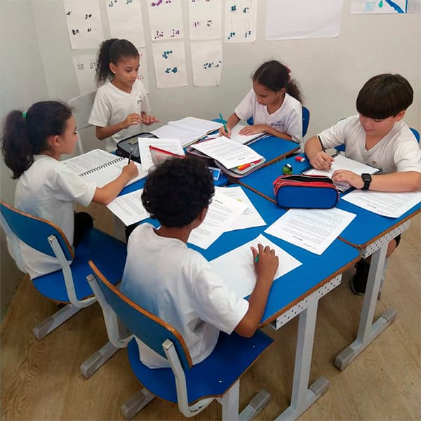 Grupo de alunos do fundamental I estudando em carteiras unidas, com folhas e cadernos abertos