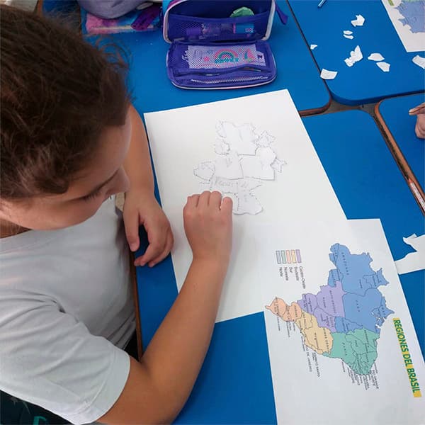 Aluna do fundamental I montando um mapa com recortes de papel, ao lado de um mapa das regiões do Brasil