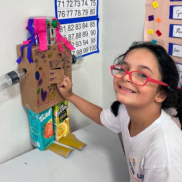 Aluna do fundamental I sorrindo ao lado do robô que construiu com caixas de papelão, embalagens e garrafas recicladas