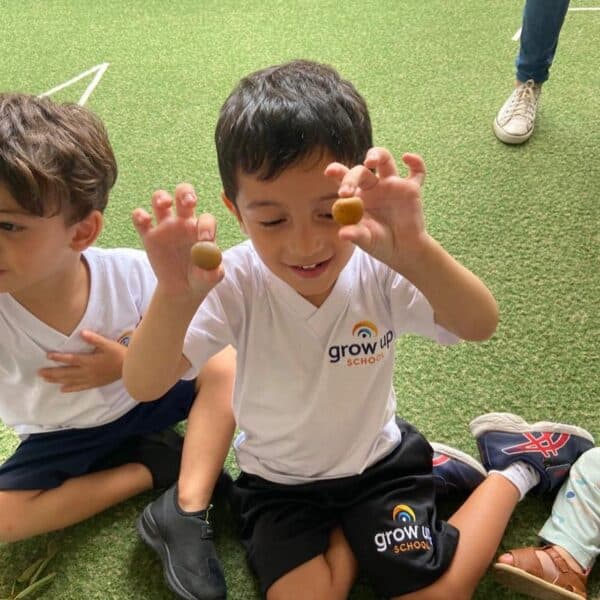 Menino da educação infantil sentado no pátio, mostrando sorridente as duas bolinhas que tem nas mãos