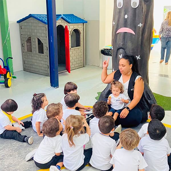 Educadora sentada junto a uma árvore decorada contando uma história para a turma da educação infantil, sentada em roda no chão